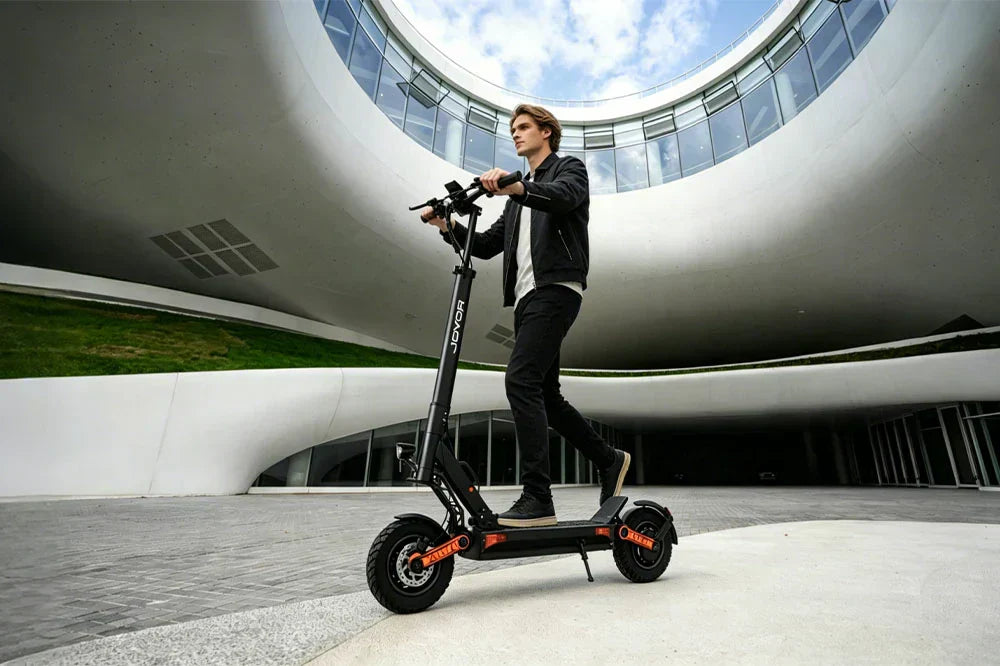 Lifestyle photo of a man riding the JOYOR S-PRO electric scooter in a modern urban plaza, showcasing its black frame, orange suspension accents, and all-terrain tires for city commuting